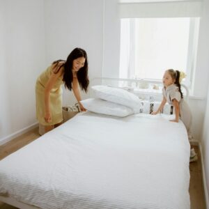 Mother and daughter smiling while making the bed in a bright, cozy bedroom.